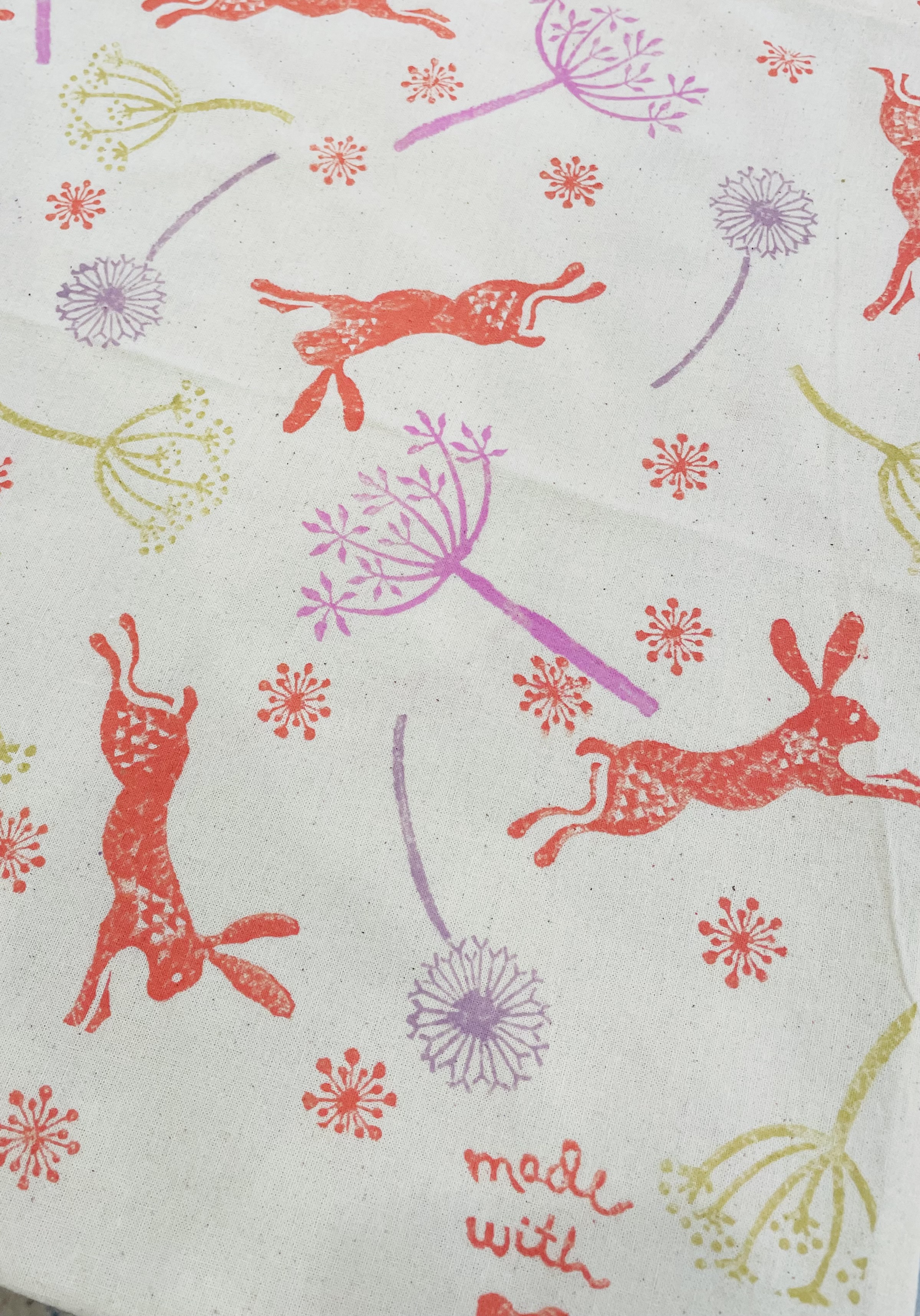A tote bag with orange leaping hares, purple and pink seed heads and orange floating seeds.